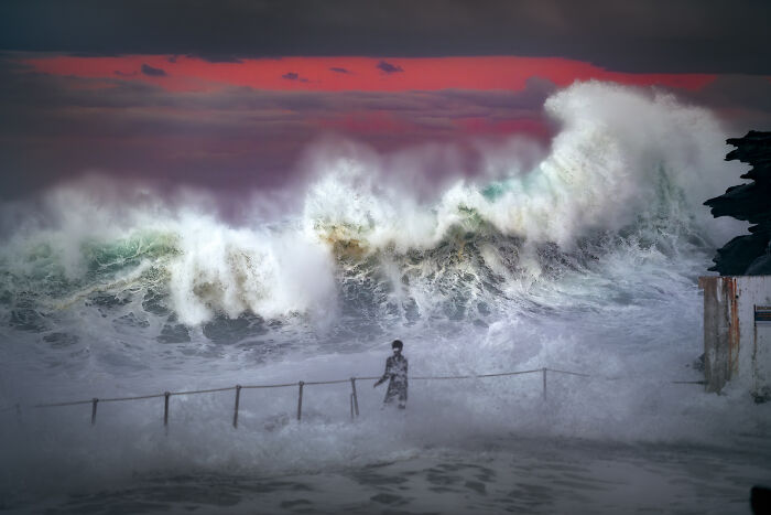 A person standing by a fence near a huge wave under a red sky, from the Ocean Photographer Of The Year Awards.