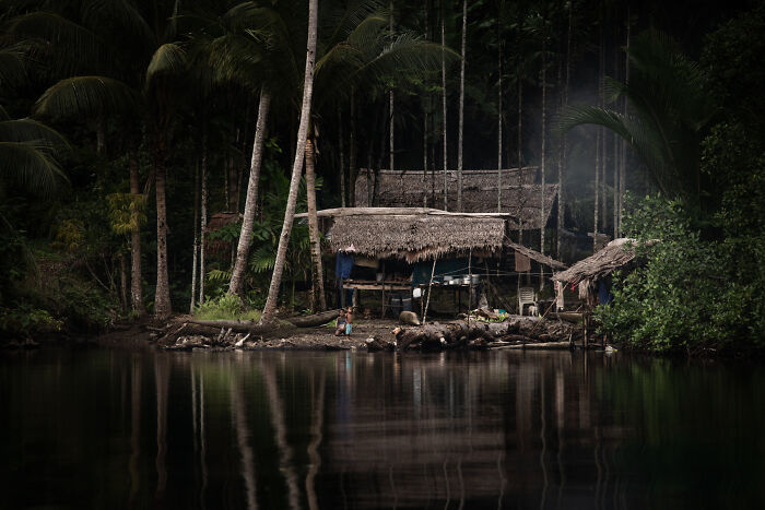 Huts with thatched roofs on a riverbank surrounded by palm trees, a stunning image from the 2026 Ocean Photographer of the Year Awards.