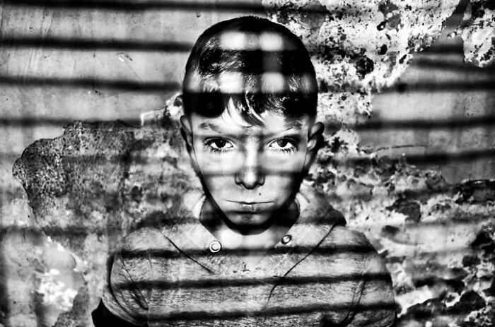 A black and white street photo of a young boy looking directly at the camera, with grid-like shadows cast across his face, depicting the intensity of childhood.