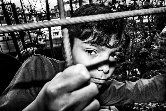A black and white street photo of a young boy's intense gaze through a blurred metal bar, focusing on the curiosity of childhood.
