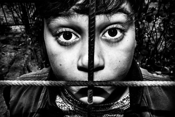 A close-up, black and white street photo of a young boy's face split by a vertical and horizontal metal bar, capturing the intensity of childhood.
