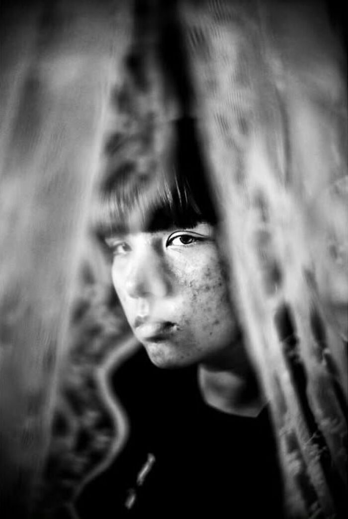 A black and white street photo by Murat Harmanlikli revealing the intensity of childhood, with a child peeking through a curtain.