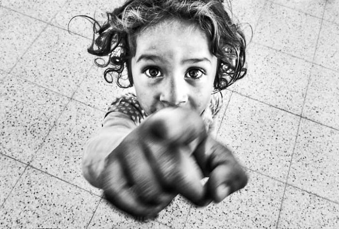 A street photo by Murat Harmanlikli shows a child with curly hair pointing directly at the camera, embodying childhood joy.