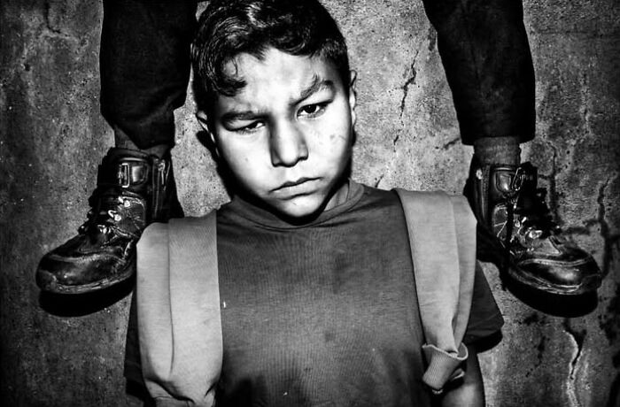 A black and white street photo capturing the intensity of childhood, featuring a young boy looking down with another person's legs above him.