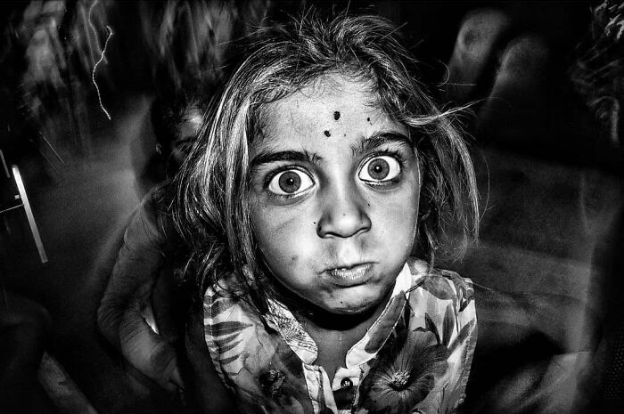A black and white street photo capturing the curiosity and intensity of childhood, showing a young girl with wide eyes and a pouting face.