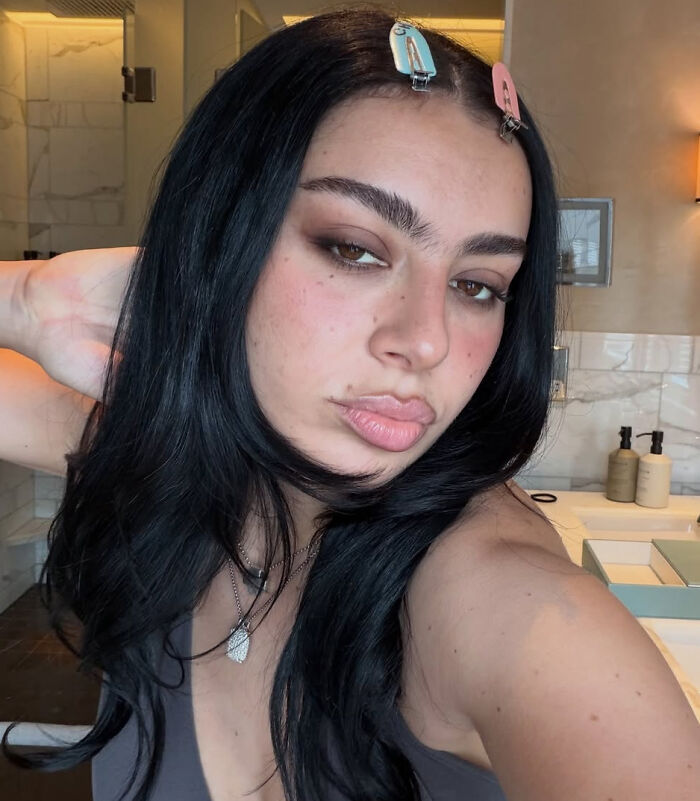 Charli XCX with dark hair and makeup, hair clips, and necklaces, representing celebrity hot takes.