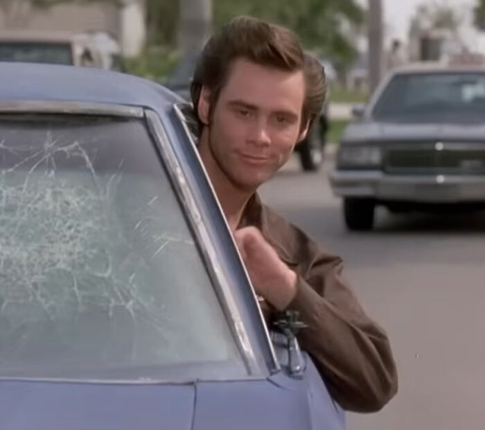 A smiling Jim Carrey peeking from a car with a broken windshield, an image related to celebrity hot takes.