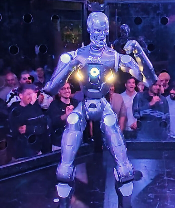 Robot flexing in cage fight arena with excited crowd watching man vs robot match