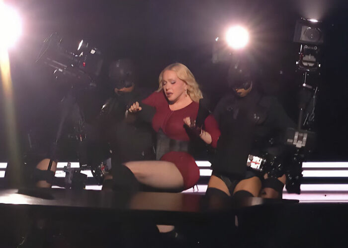Madonna seated on stage in red outfit supported by dancers amid bright lights Madonna seated on stage in red outfit supported by dancers amid bright lights