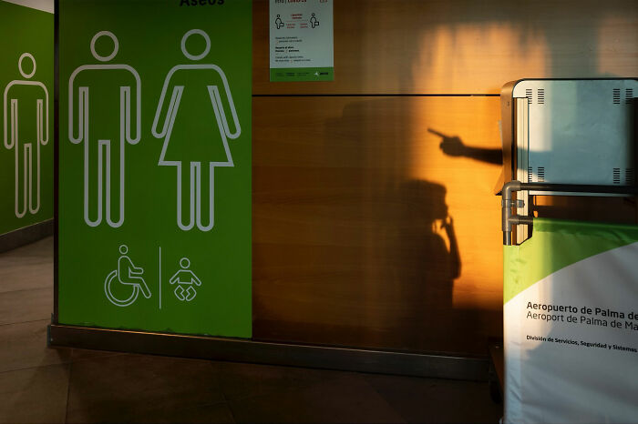 A long shadow pointing towards a restroom sign with universal access symbols, an odd coincidence captured by a street photographer.