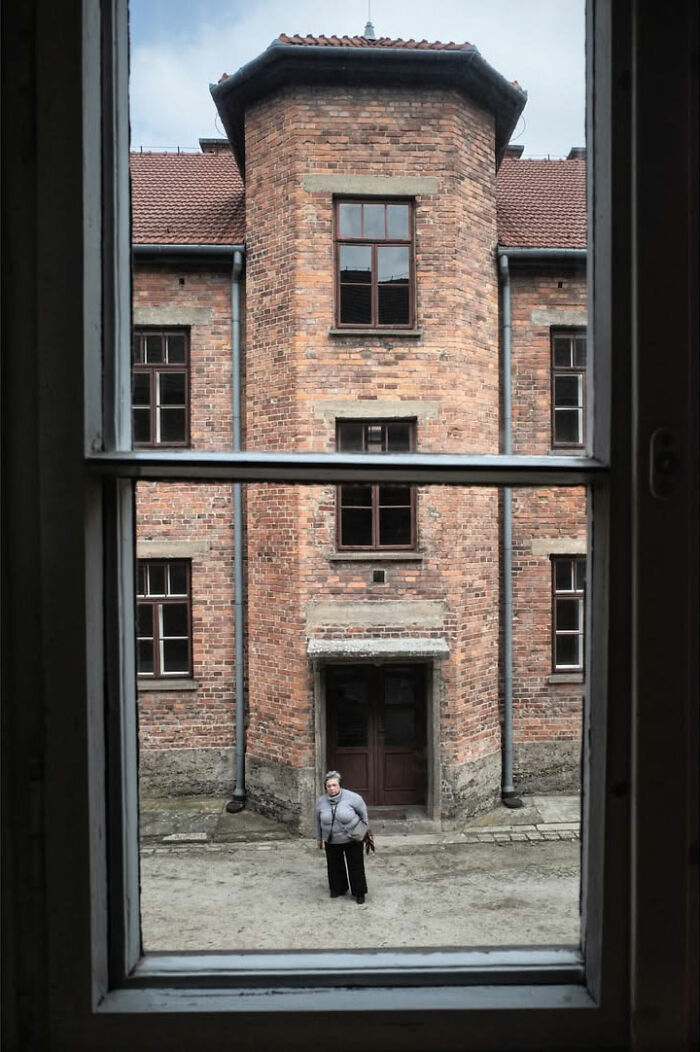 A person standing in a courtyard viewed through a window, framed by an old brick building, a scene captured by a street photographer.