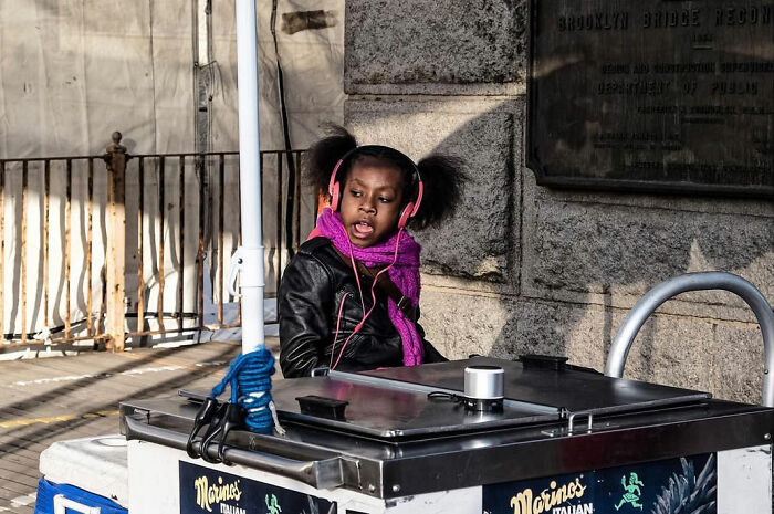A young girl in pink headphones and a purple scarf singing at a street vendor's cart, an odd coincidence captured by a street photographer.