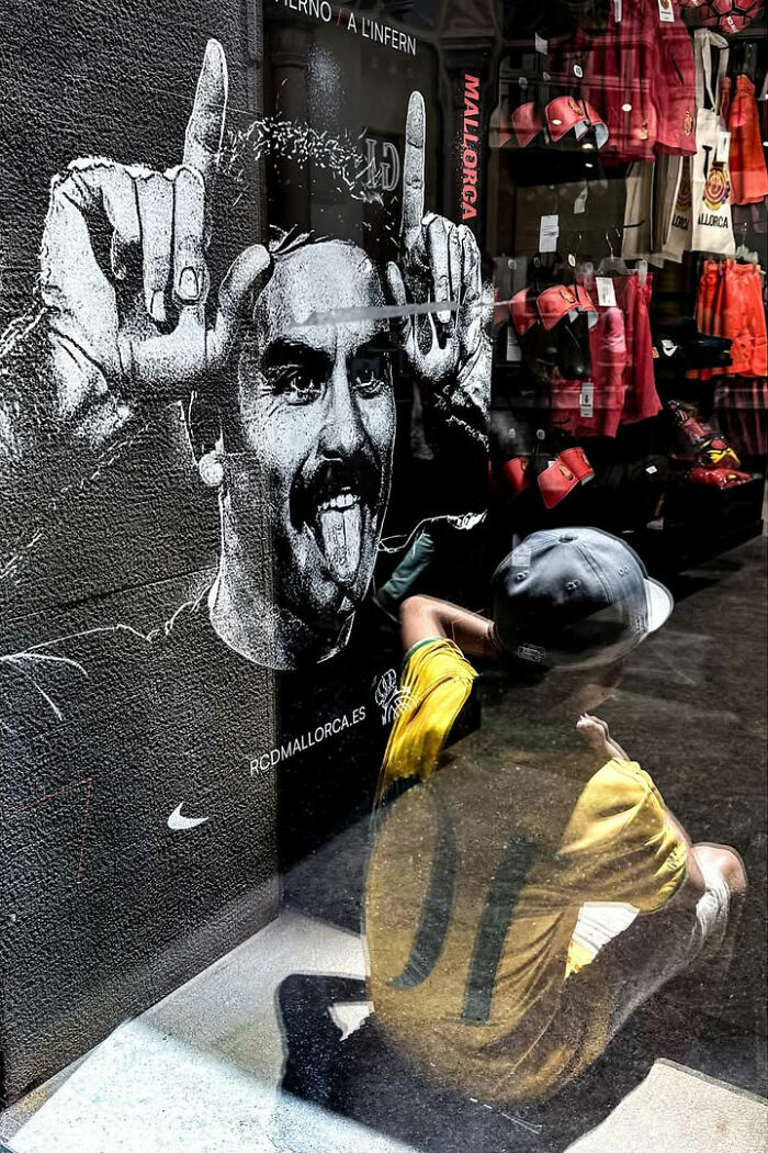 A street photographer captured an odd coincidence: a boy looking at a window with a graffiti of a man making a funny face, which is reflected in the glass.