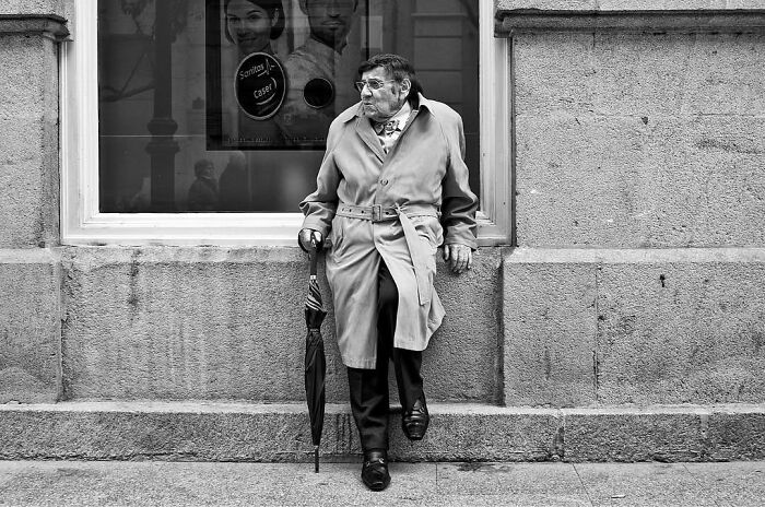 A street photographer captures an old man in a trench coat leaning against a wall with an umbrella, an odd street scene.