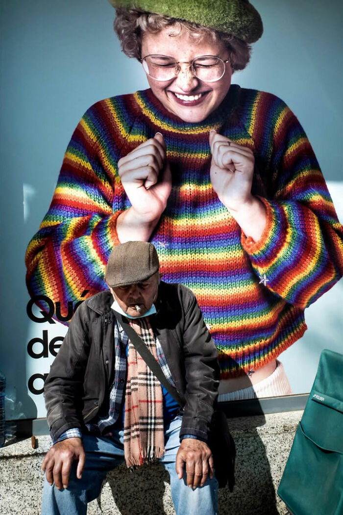 A street photographer captures an old man smoking, sitting in front of a colorful advertisement, an odd street scene.