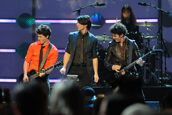 A band of celebrities performing live on stage with guitars and a drum set, capturing brutal hot takes.