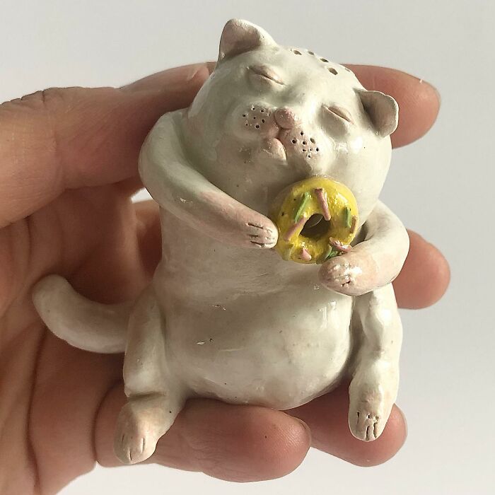 A hand holding a white cat weird ceramic sculpture eating a tiny donut, ideal for quirky home decor.