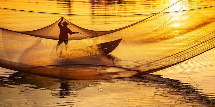 Remarkable travel photos of a fisherman casting a large net at sunset, creating a golden glow on the water.