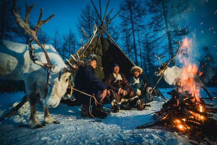 Three people in traditional winter attire sit by a campfire with reindeer in a snowy forest, a remarkable travel photo.