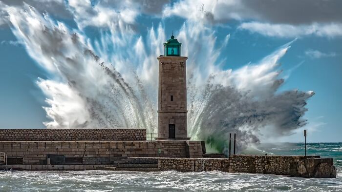 A lighthouse endures a powerful wave crashing behind it under a dramatic sky, a remarkable travel photo.