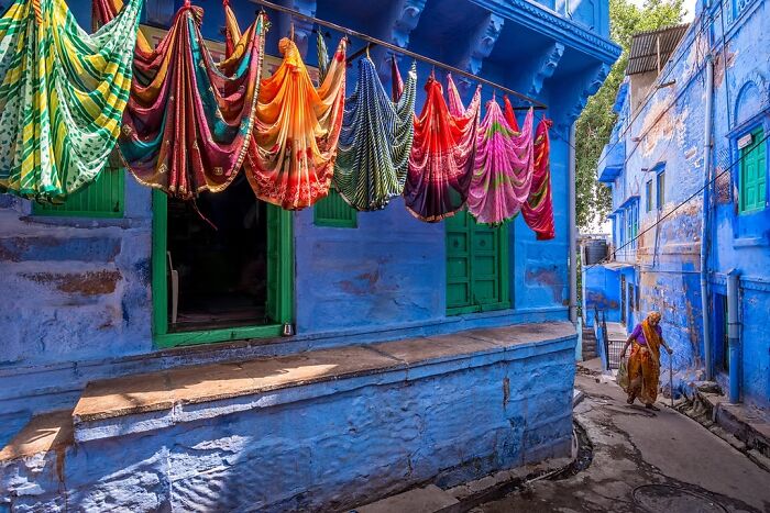 Colorful textiles hang to dry against a vibrant blue building in a narrow alley, a remarkable travel photo.