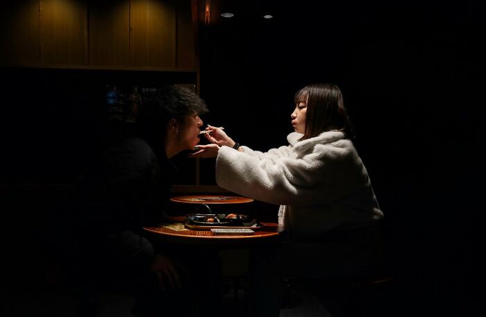 A woman feeding a man in a dimly lit restaurant, a candid street photo for URBAN Photo Awards.