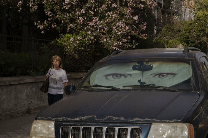 A woman walks past a car with a sunshade showing large eyes, a candid street photo for URBAN Photo Awards.