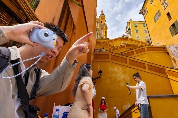 A person points while holding a light blue instant camera, with others on yellow stairs in the background, a candid street photo for the URBAN Photo Awards.