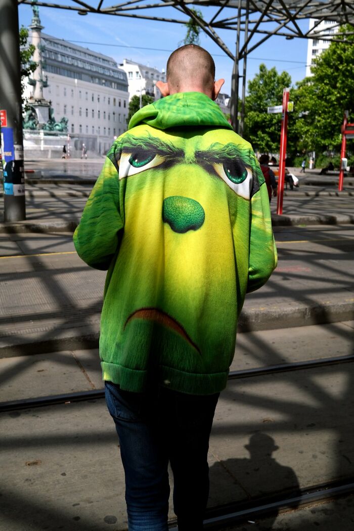 A person from behind wears a green Grinch-like hoodie with pointy ears, walking on a city street, a candid street photo for the URBAN Photo Awards.