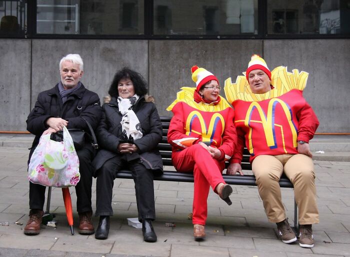 Two people dressed in McDonald's french fries costumes sit on a bench with two elderly people, a candid street photo from the URBAN Photo Awards.