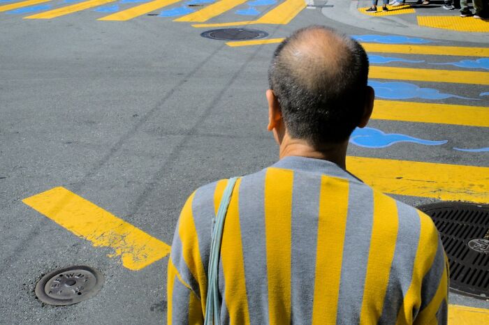 A man with a bald head and a yellow and grey striped sweater stands looking at a street crossing with yellow and blue markings, part of the URBAN Photo Awards.