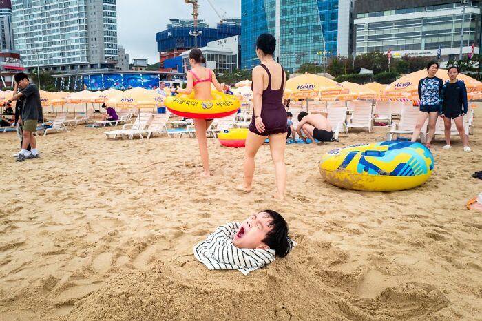 A child buried in sand on a crowded beach with buildings in the background, a street photo from URBAN Photo Awards.