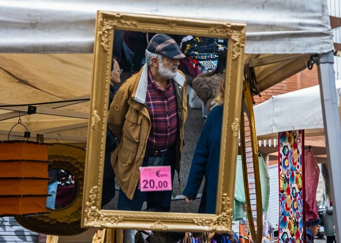 A man reflected in an ornate gold frame hanging at an outdoor market, a candid street photo for URBAN Photo Awards.