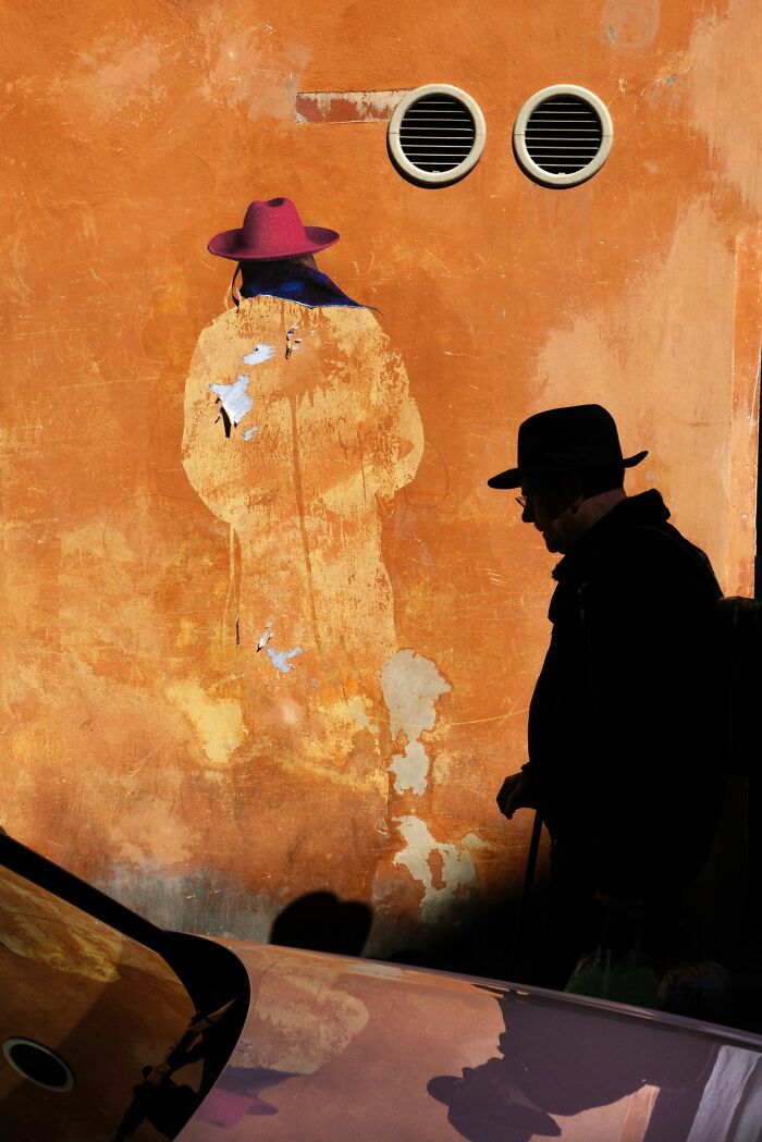 A man in a fedora walking past an old orange wall with a faded outline of a person wearing a hat, a candid street photo for the URBAN Photo Awards.