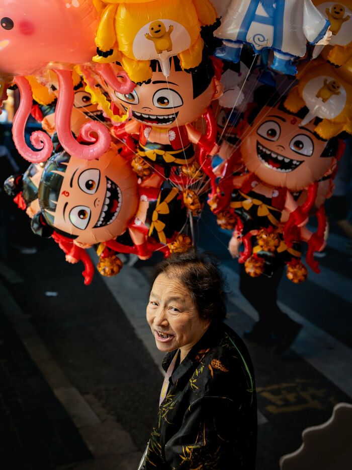 A smiling woman looking up, surrounded by colorful cartoon-character balloons, a candid street photo for the URBAN Photo Awards.