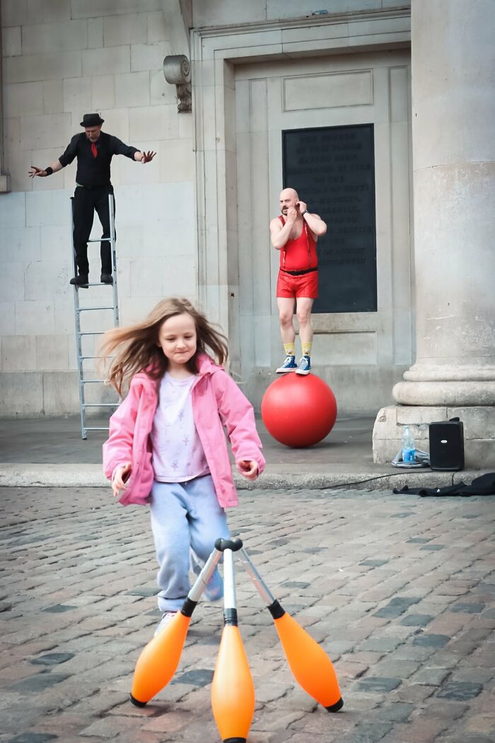 A lively street performance featuring a child, a juggler on a ladder, and another performer on a red ball, from the URBAN Photo Awards.