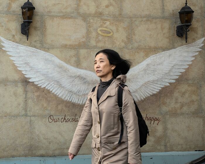 A woman walking past a wall mural with painted angel wings and a halo, a candid street photo.