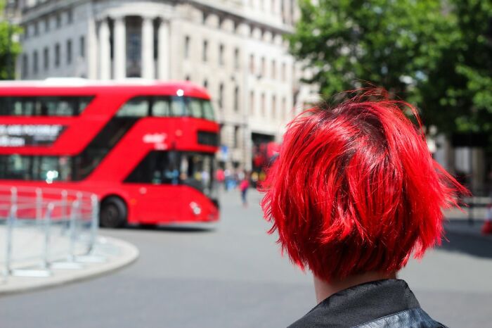 Person with bright red hair facing away, with a red double-decker bus in the background of a street photo, part of URBAN Photo Awards.