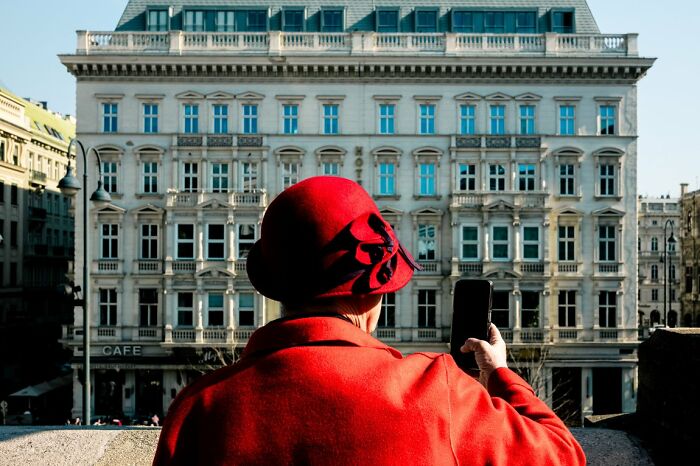 Woman in a red coat and hat taking a photo of a grand building, a candid street photo from URBAN Photo Awards.
