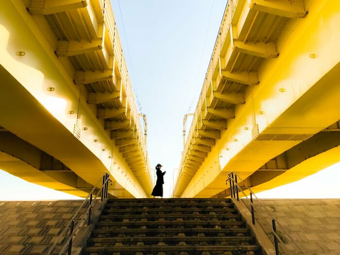 A woman stands under a bright yellow bridge, captured in a candid street photo from the URBAN Photo Awards.