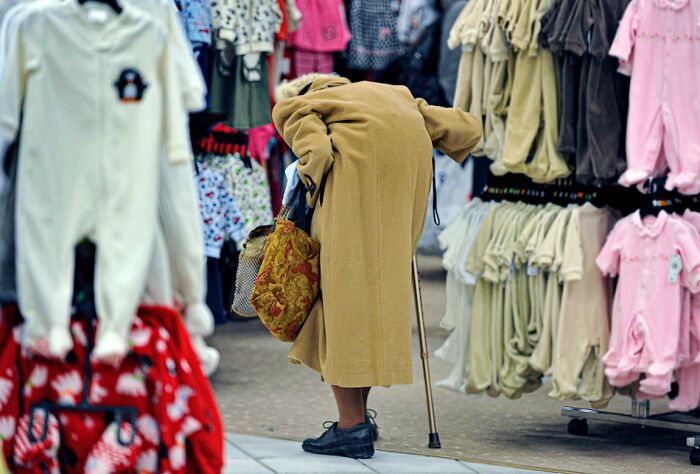 Candid Street Photos of an elderly person with a cane bent over in a market stall among baby clothes.
