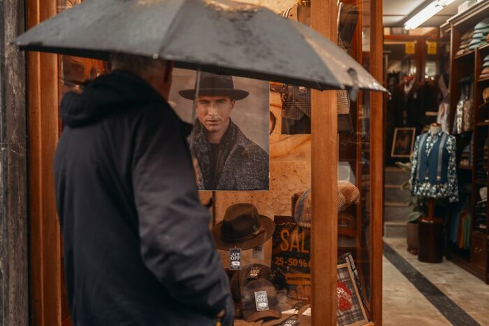 A person with an umbrella looks into a hat shop window, a candid street photo showcasing urban life for the URBAN Photo Awards.