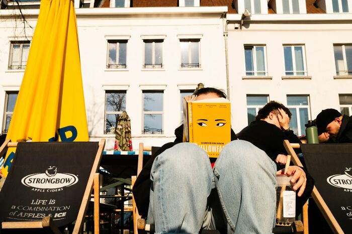 A person reads a yellow book with eyes depicted on it, relaxing outdoors in a candid street photo from URBAN Photo Awards.