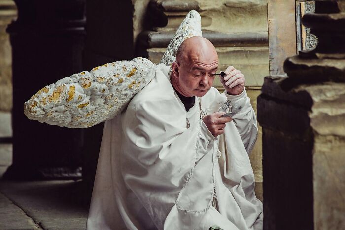 Candid Street Photos of a bald man with white robes and large, textured wings, applying something to his eye.