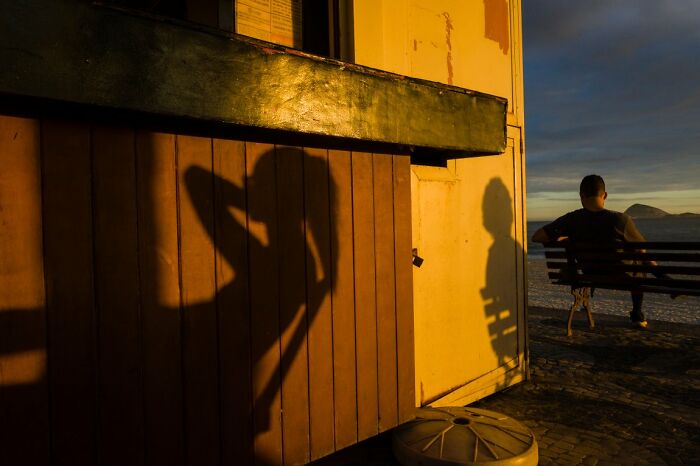 Candid Street Photos of a man sitting on a bench with long shadows cast by a building at sunset.