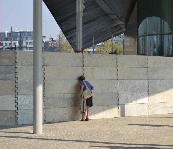 Candid Street Photos of a person in a blue shirt and shorts looking through a small opening in a concrete wall.