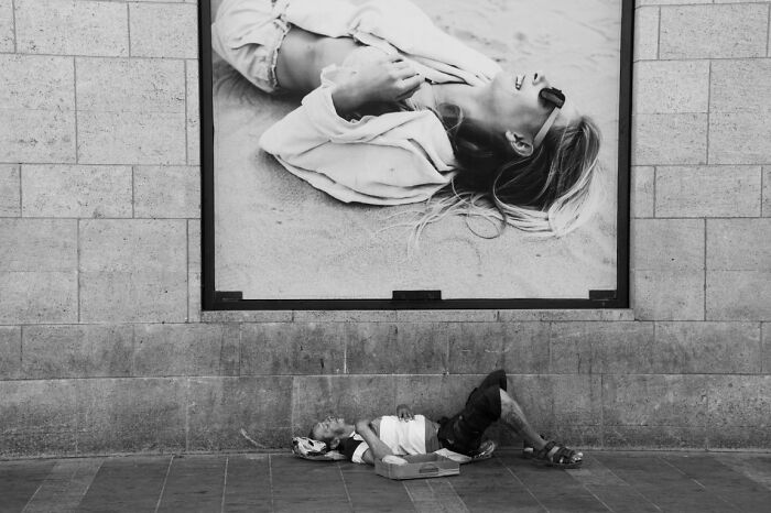A candid street photo of a person sleeping on the ground below a large advertisement for the URBAN Photo Awards.