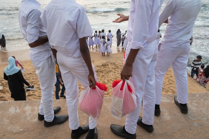 Candid street photo of men in white clothes holding red bags on a beach, with people near the water for the URBAN Photo Awards.