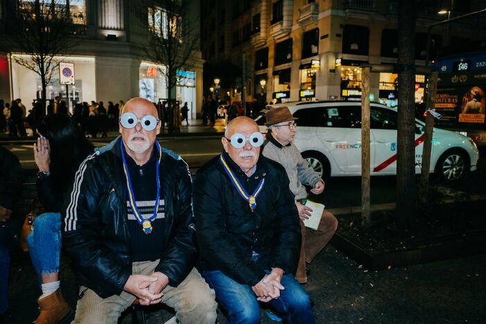 Two men with googly-eye glasses sitting on a bench at night, a candid street photo for URBAN Photo Awards.