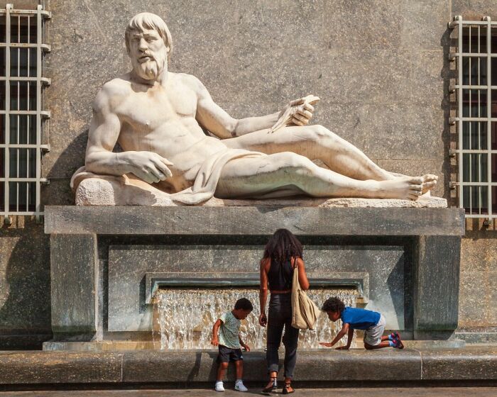A candid street photo of a large stone statue with a woman and two children below, part of the URBAN Photo Awards.